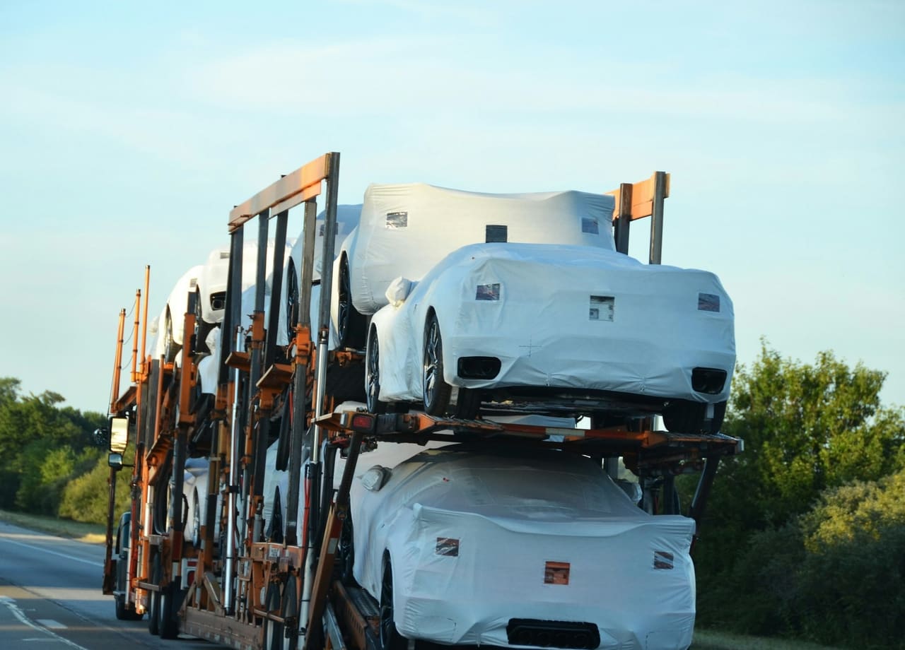 A loaded auto-transport carrier on a highway, sports cars under white transit covers strapped across its upper and lower decks.
