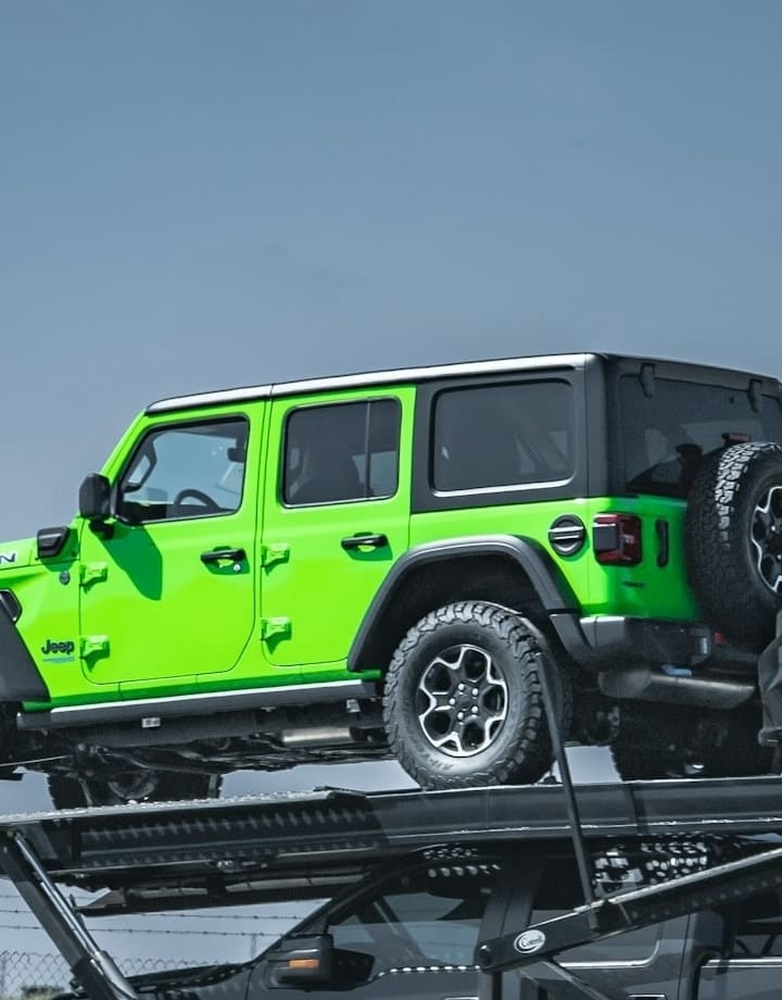 A four-door off-roader standing on the upper deck of a car carrier, wheels seated on the steel grating against clear sky.