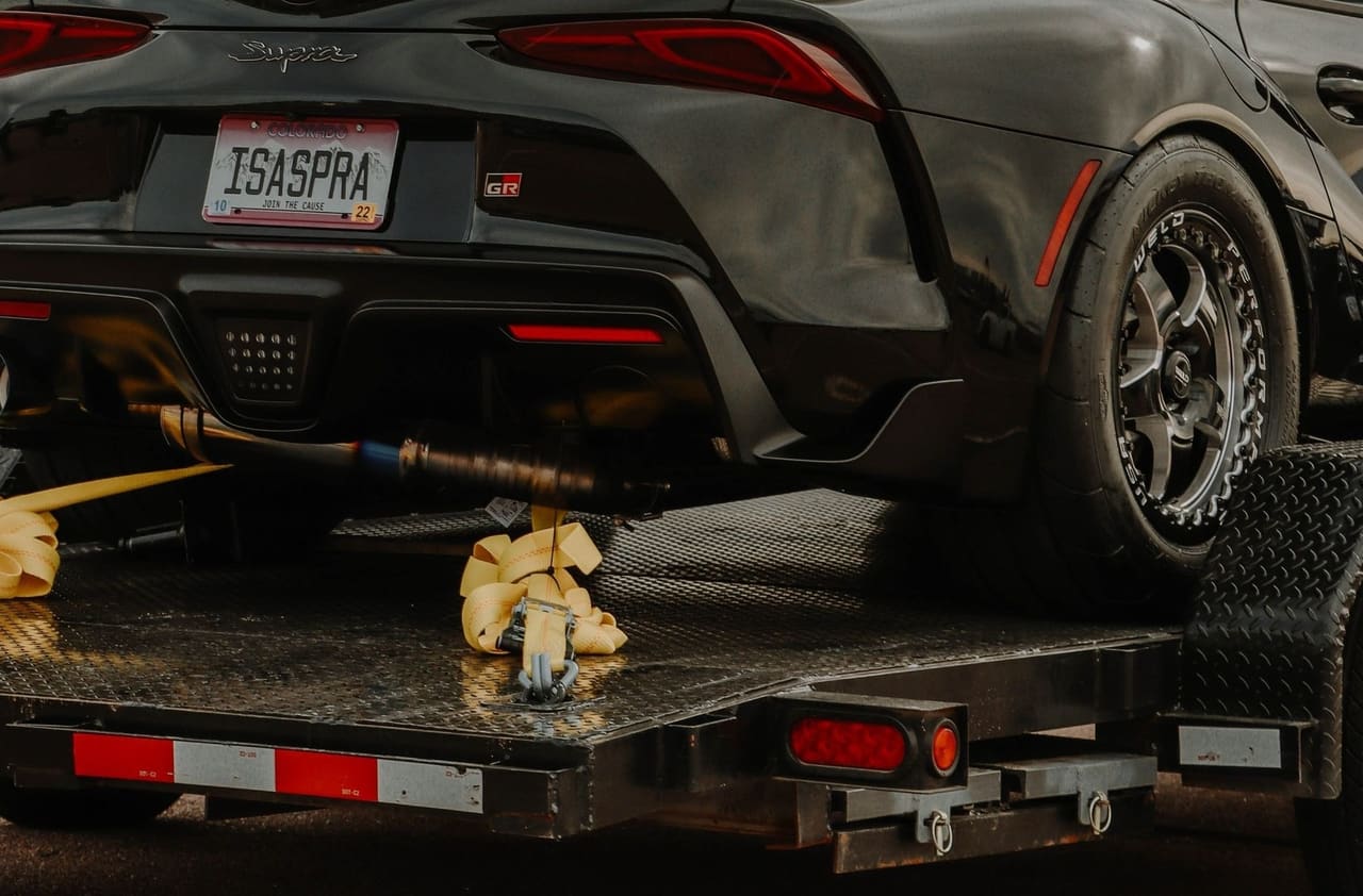 A wheel strap ratcheted down to the deck rail beside a loaded vehicle, the trailer's marker tape below.